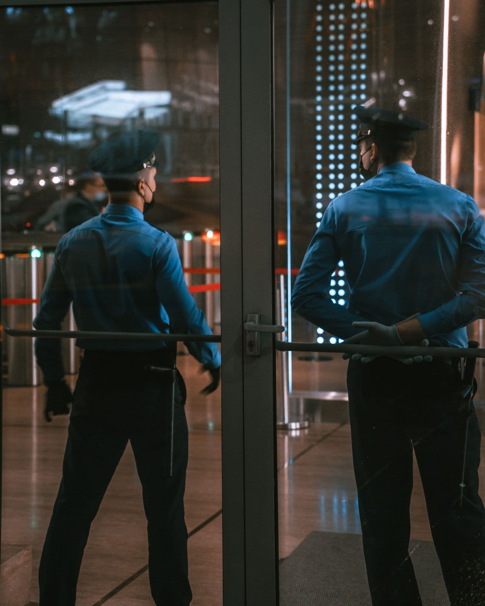 Dos guardias de seguridad uniformados vigilando de noche junto a la puerta de cristal de un edificio.