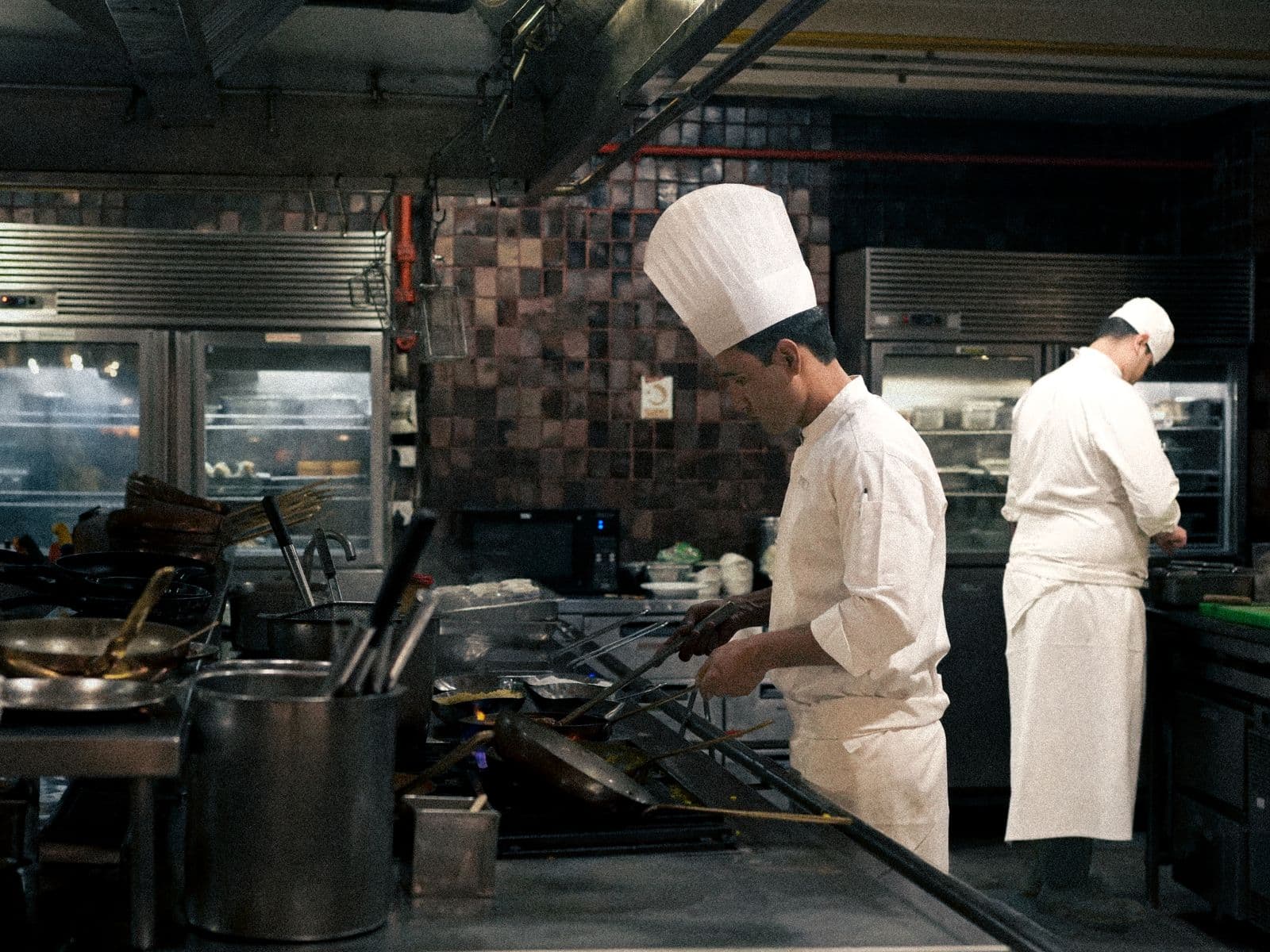 Dos cocineros con gorro y uniforme trabajando junto a los fogones en plena cocina profesional de un restaurante.