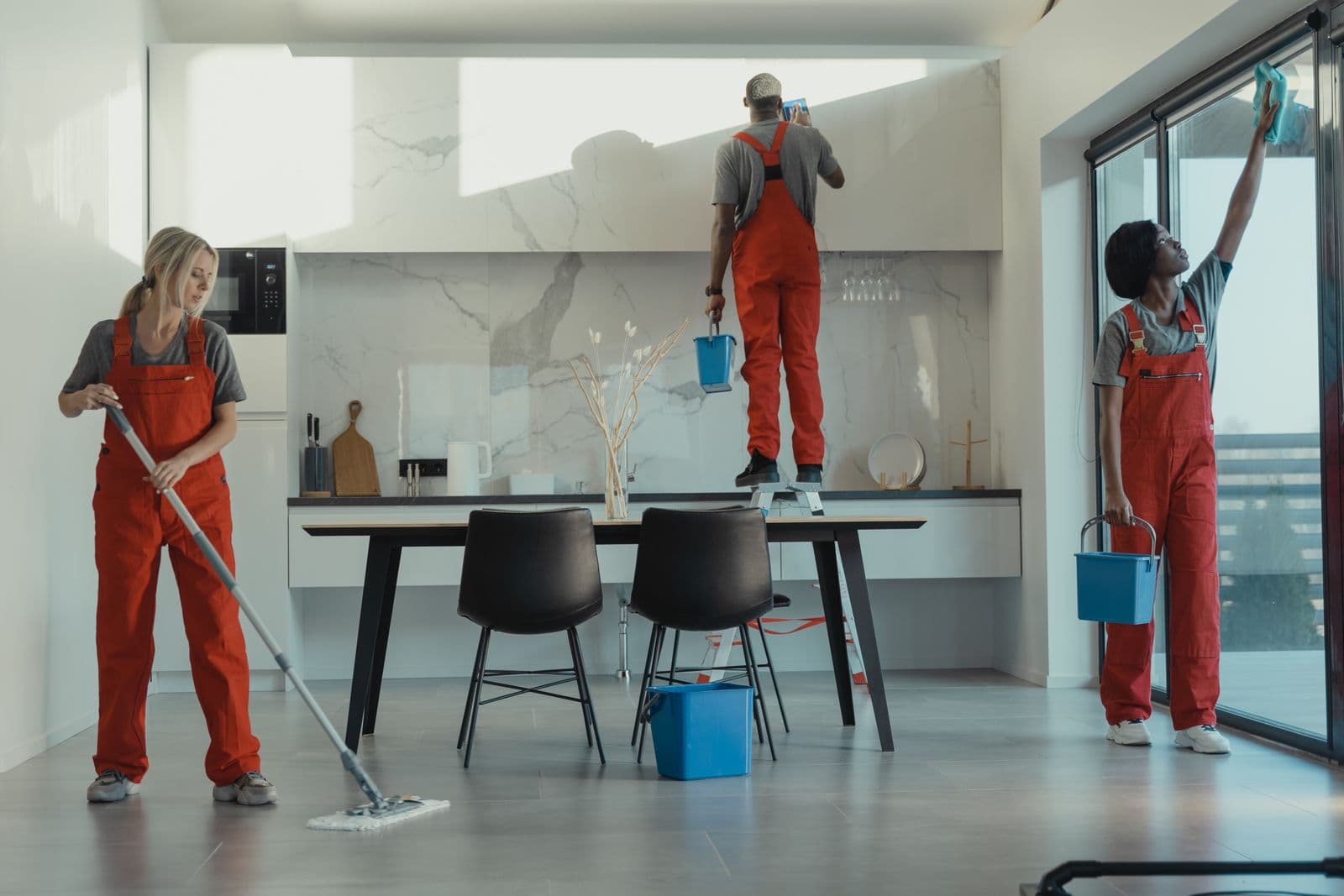 Un equipo de limpieza profesional con mono de trabajo naranja friega el suelo, limpia una superficie en altura y abrillanta un cristal en un interior moderno.